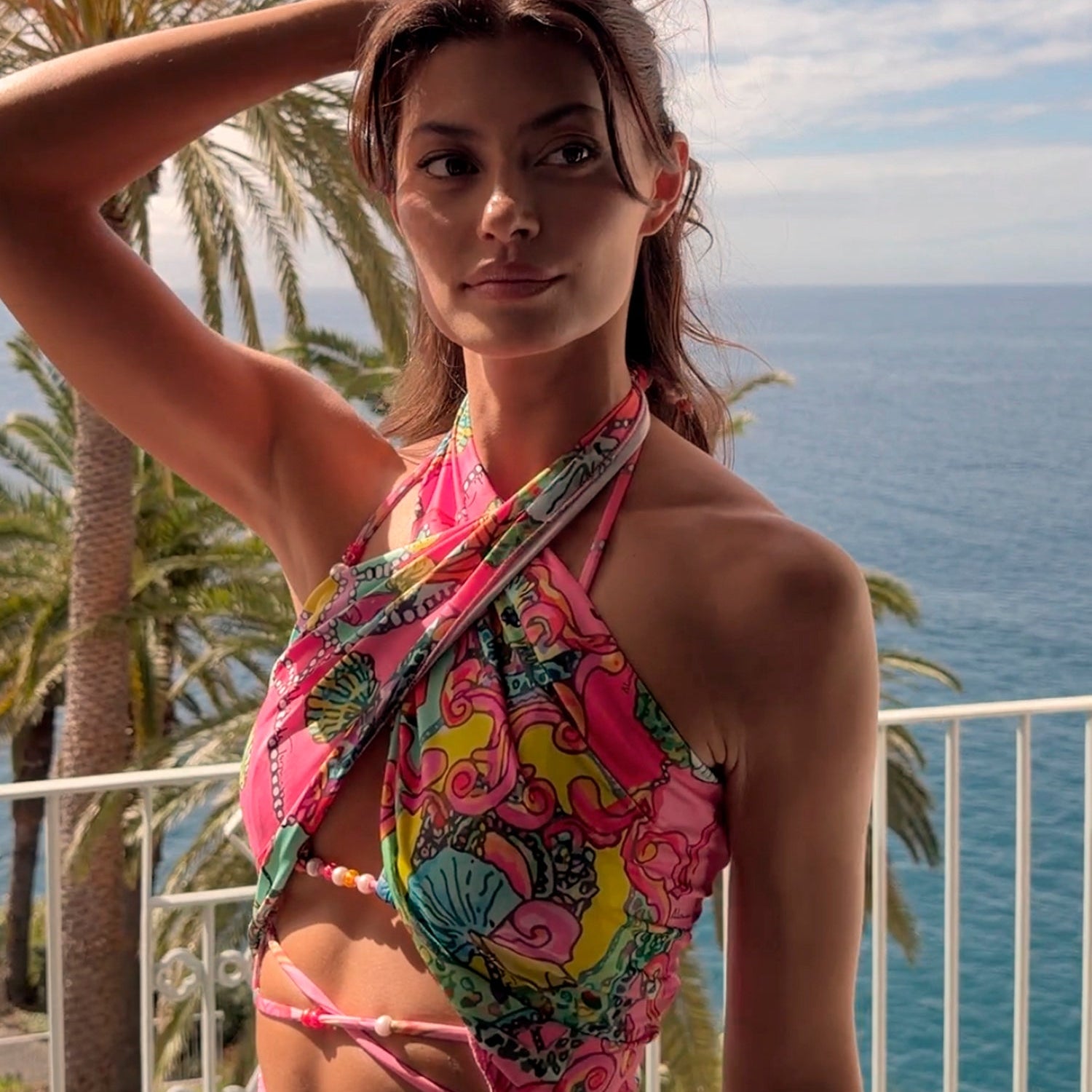 Coralia 3-in-1 worn as scarf top close-up on model — coral pink multicolour floral crochet piece wrapped and tied as a halter top, worn on a sea-view balcony with palm trees, handcrafted in Portugal by Alameda Turquesa