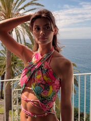 Coralia 3-in-1 worn as scarf top close-up on model — coral pink multicolour floral crochet piece wrapped and tied as a halter top, worn on a sea-view balcony with palm trees, handcrafted in Portugal by Alameda Turquesa