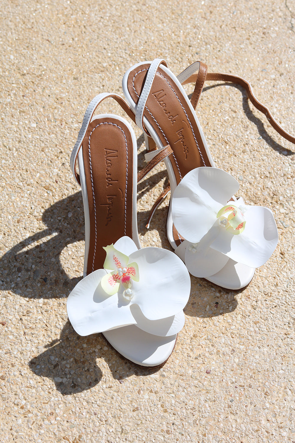 Fiona white heels flat lay on sand — white leather ankle-strap stiletto heels with handcrafted white orchid embellishments and Alameda Turquesa leather insole, handcrafted in Portugal by Alameda Turquesa