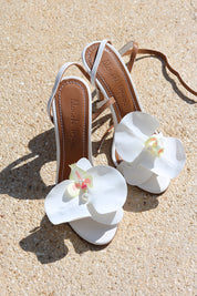 Fiona white heels flat lay on sand — white leather ankle-strap stiletto heels with handcrafted white orchid embellishments and Alameda Turquesa leather insole, handcrafted in Portugal by Alameda Turquesa