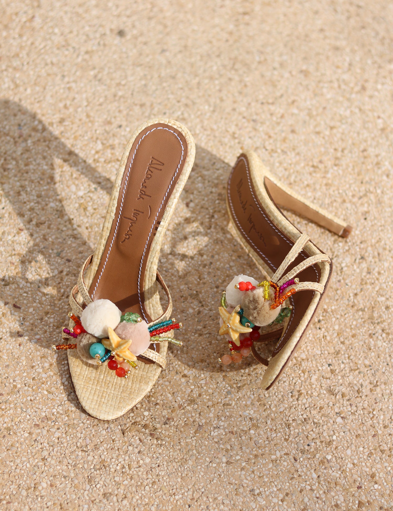 Isla raffia mules flat lay — natural raffia straps with hand-applied starfish, shell and multicolour bead embellishments on sand, luxury mules by Alameda Turquesa Portugal
