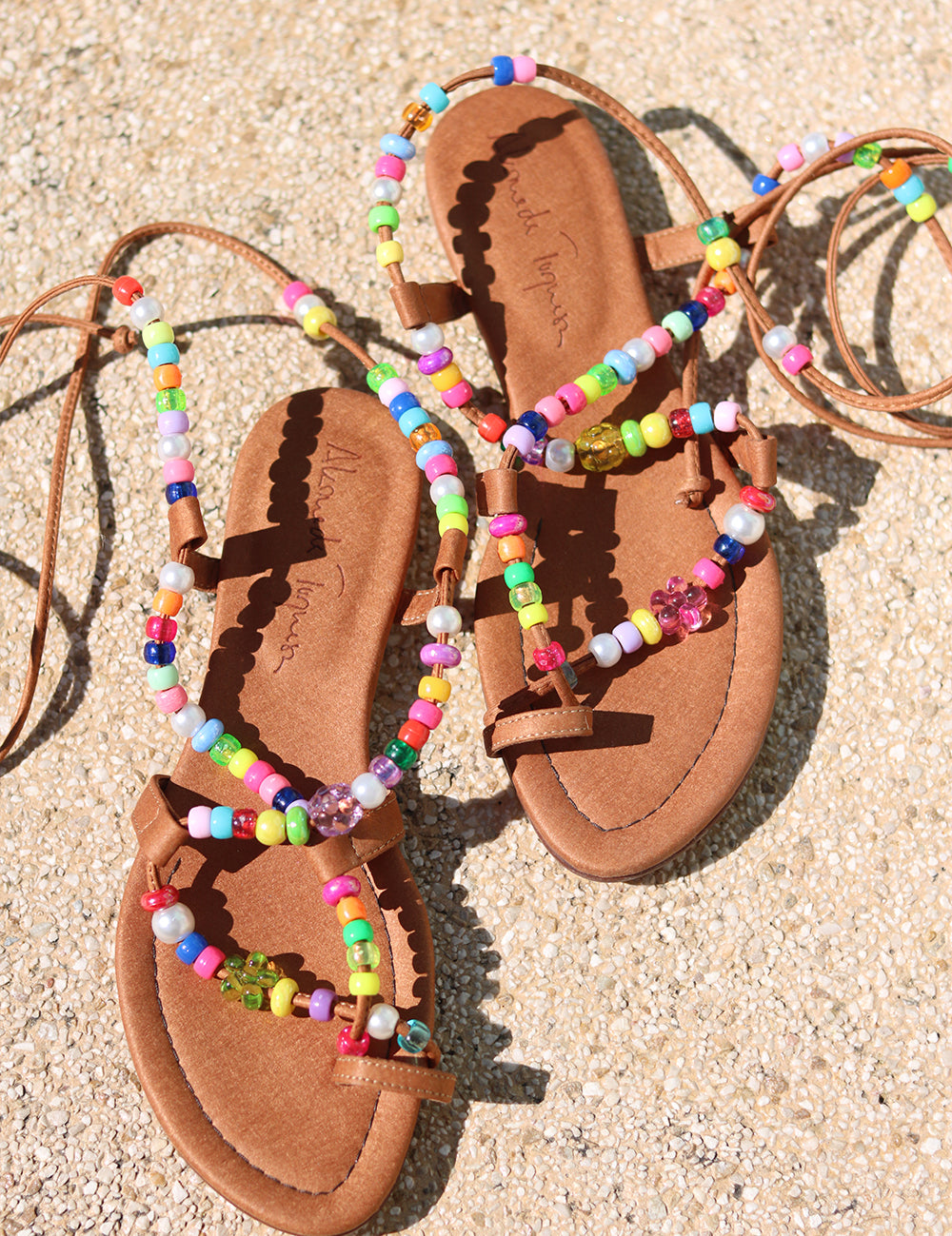 Alegria beaded sandals flat lay — tan Italian leather strappy flat sandals with multicolour handcrafted bead embellishments and long tie laces, pair styled overhead on sandy ground, handcrafted in Portugal by Alameda Turquesa