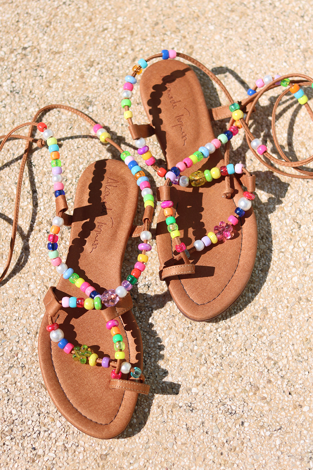 Alegria beaded sandals flat lay — tan Italian leather strappy flat sandals with multicolour handcrafted bead embellishments and long tie laces, pair styled overhead on sandy ground, handcrafted in Portugal by Alameda Turquesa