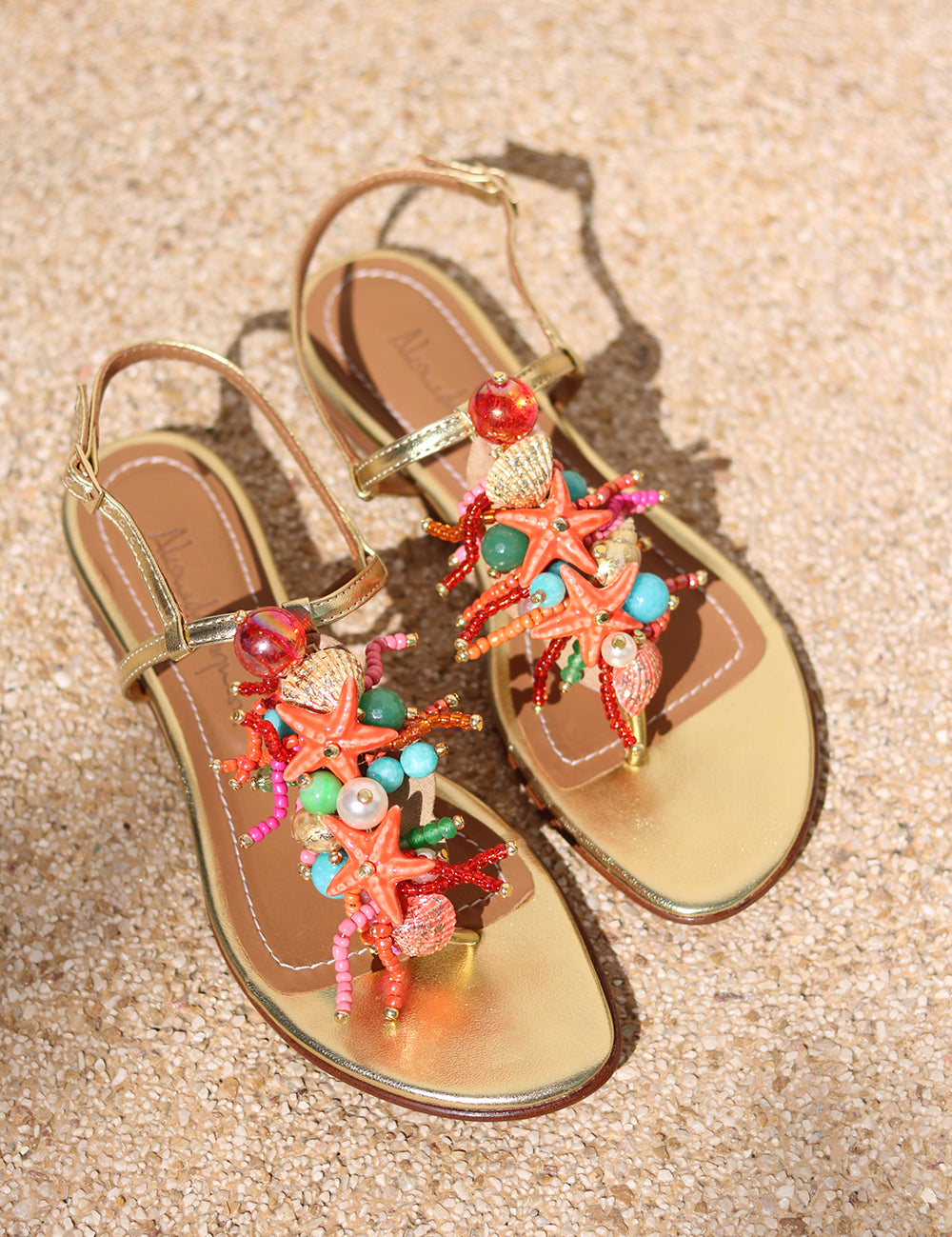 Atlântica sandal flat lay on sand — gold leather toe-post flat sandal with hand-applied coral starfish, natural shells, pearls, turquoise stones and multicolour bead embellishments, Alameda Turquesa leather insole, handcrafted in Portugal by Alameda Turquesa