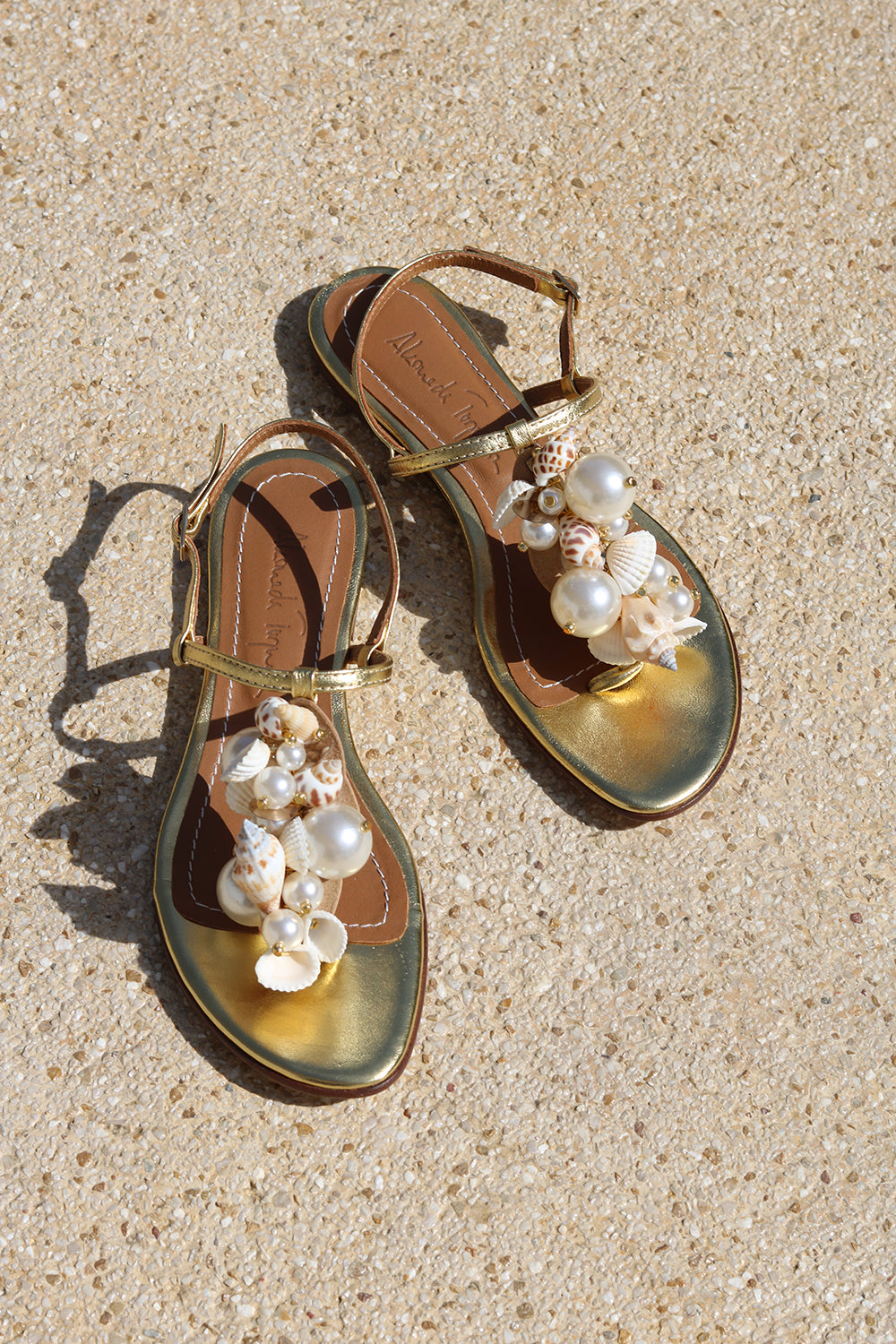 Belize sandals flat lay on sand — gold leather toe-post flat sandals with hand-applied oversized pearls and natural shell embellishments, Alameda Turquesa leather insole, handcrafted in Portugal by Alameda Turquesa