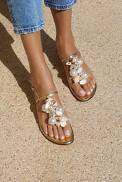 Belize sandals close front view on both feet — gold leather toe-post flat sandals with hand-applied oversized pearls, natural shells and gold bead embellishments with ankle strap, styled with denim jeans on sand, handcrafted in Portugal by Alameda Turquesa
