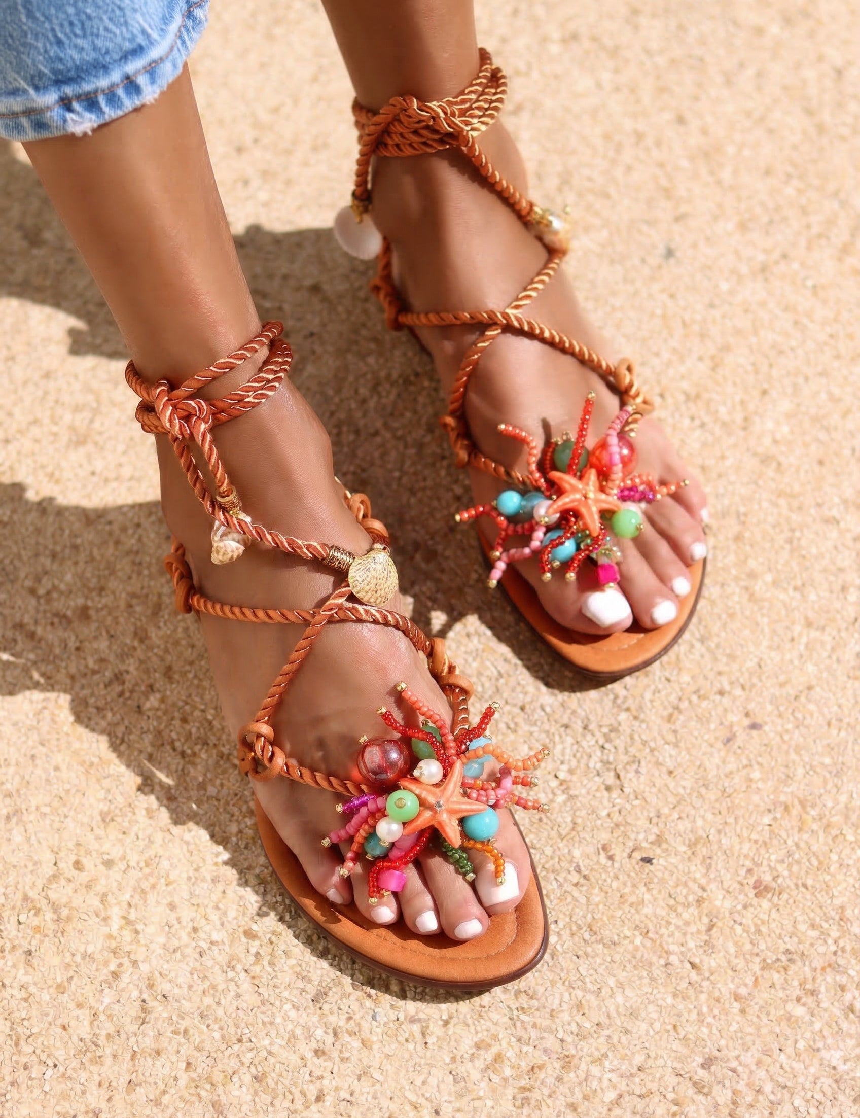 Caribe sandals front view on both feet — tan leather toe-post flat sandals with hand-applied coral starfish and multicolour bead embellishments, twisted rope lace-up ankle wraps with gold coin charms, styled with denim jeans on sand, handcrafted in Portugal by Alameda Turquesa