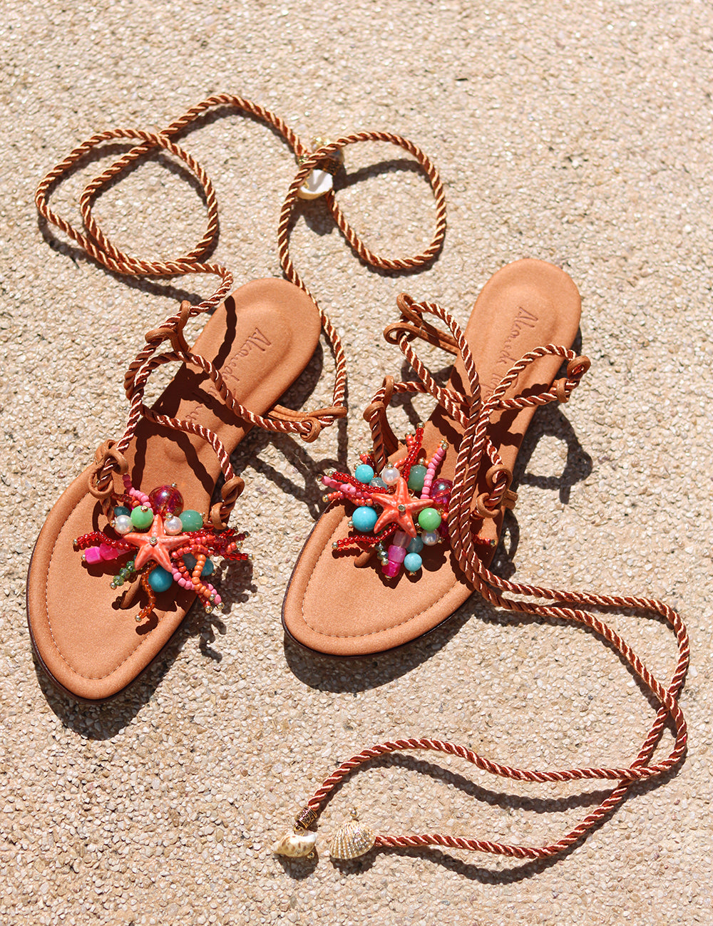 Caribe sandals flat lay on sand — tan leather toe-post flat sandals with hand-applied coral starfish and multicolour bead embellishments, twisted rope lace-up ankle wraps with shell charm tips, Alameda Turquesa leather insole, handcrafted in Portugal by Alameda Turquesa