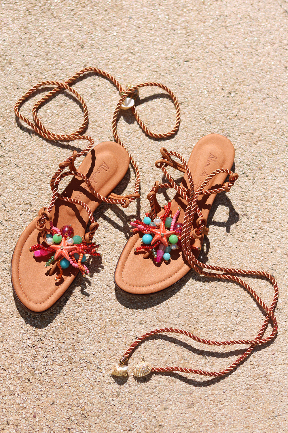 Caribe sandals flat lay on sand — tan leather toe-post flat sandals with hand-applied coral starfish and multicolour bead embellishments, twisted rope lace-up ankle wraps with shell charm tips, Alameda Turquesa leather insole, handcrafted in Portugal by Alameda Turquesa