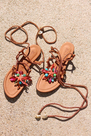 Caribe sandals flat lay on sand — tan leather toe-post flat sandals with hand-applied coral starfish and multicolour bead embellishments, twisted rope lace-up ankle wraps with shell charm tips, Alameda Turquesa leather insole, handcrafted in Portugal by Alameda Turquesa