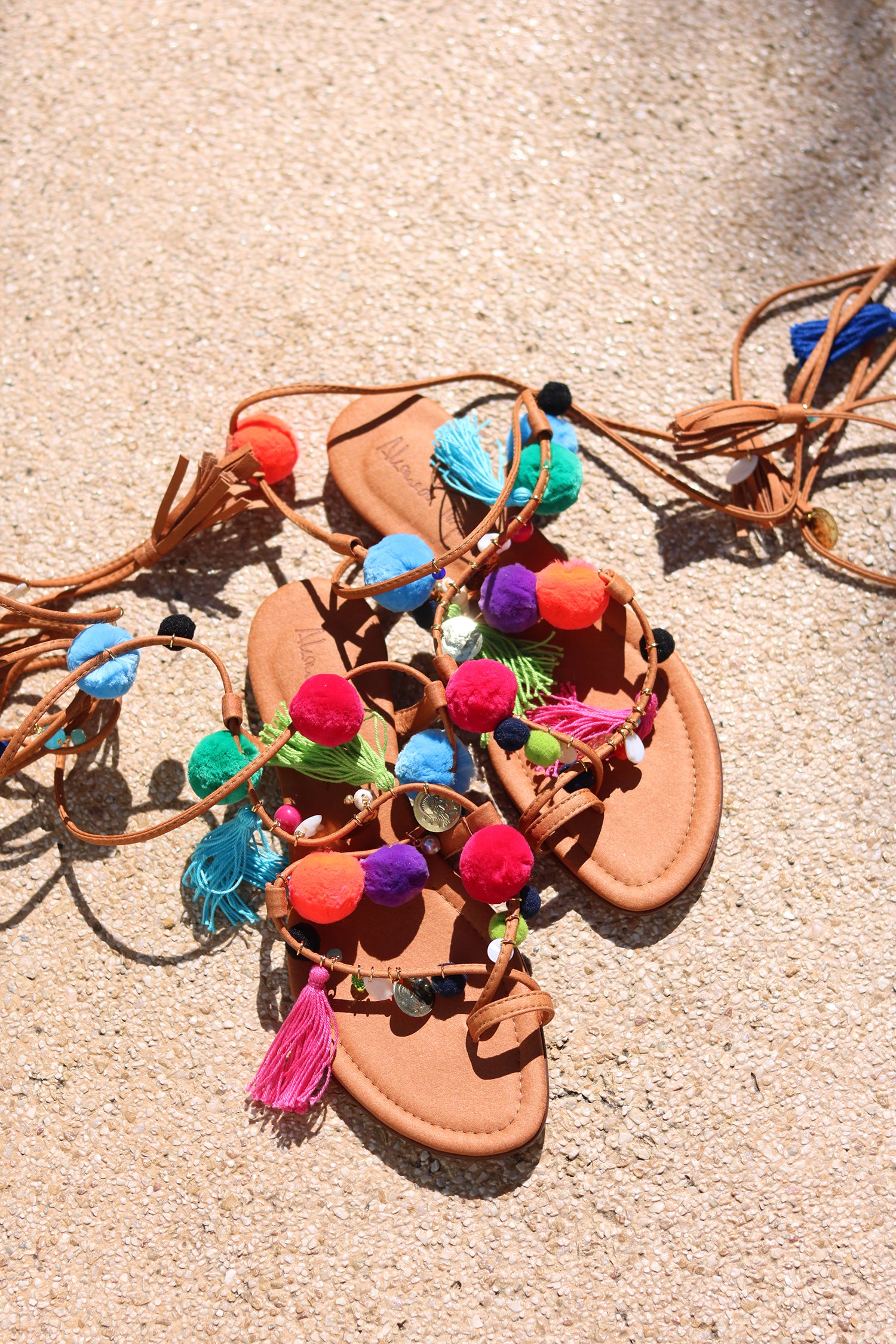 Matisse sandal flat lay on sand — tan leather toe-post flat sandal with hand-applied multicolour pom poms, silk tassels and gold coin charms, with suede lace-up ankle wrap ties, handcrafted in Portugal by Alameda Turquesa