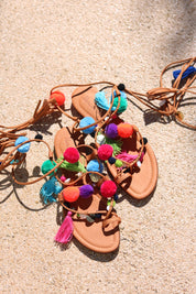Matisse sandal flat lay on sand — tan leather toe-post flat sandal with hand-applied multicolour pom poms, silk tassels and gold coin charms, with suede lace-up ankle wrap ties, handcrafted in Portugal by Alameda Turquesa