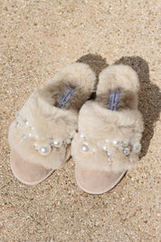 Pinctada Vanilla Pearls shearling mules flat lay — pair of ivory shearling mules with handcrafted clustered white pearl and crystal embellishments on sand suede, styled overhead on sandy ground, handcrafted in Portugal by Alameda Turquesa