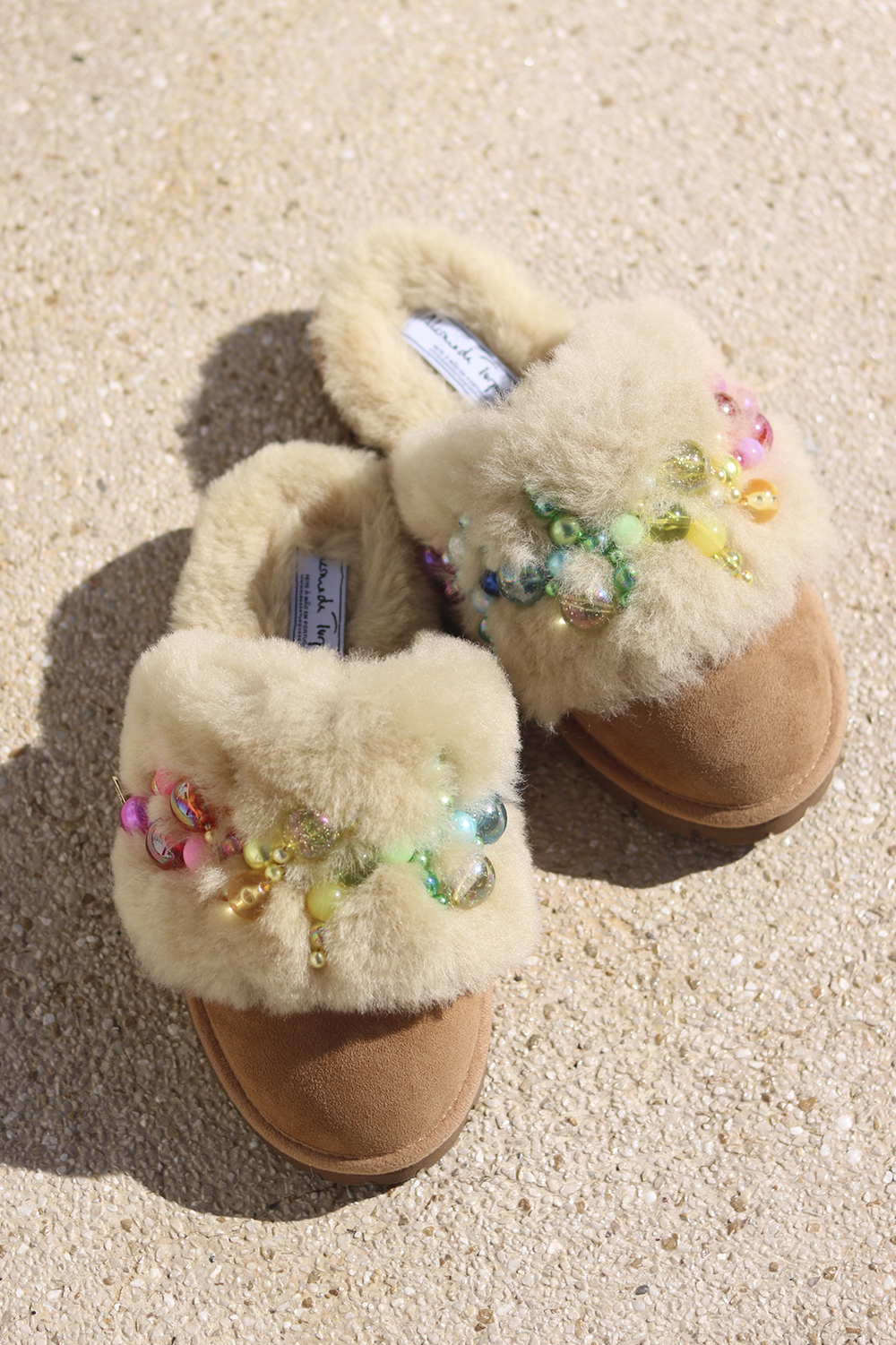 Pinctada Rainbow shearling mules flat lay — pair of cream shearling mules with handcrafted rainbow pearl and bead embellishments in pink, orange, green and yellow on caramel suede, styled overhead on sandy ground, handcrafted in Portugal by Alameda Turquesa
