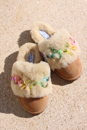 Pinctada Rainbow shearling mules flat lay — pair of cream shearling mules with handcrafted rainbow pearl and bead embellishments in pink, orange, green and yellow on caramel suede, styled overhead on sandy ground, handcrafted in Portugal by Alameda Turquesa