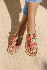 Atlântica sandals close front view on both feet — gold leather toe-post flat sandals with hand-applied coral starfish, natural shells, pearls, turquoise stones and multicolour bead embellishments, styled with denim shorts in sunlight on sand, handcrafted in Portugal by Alameda Turquesa