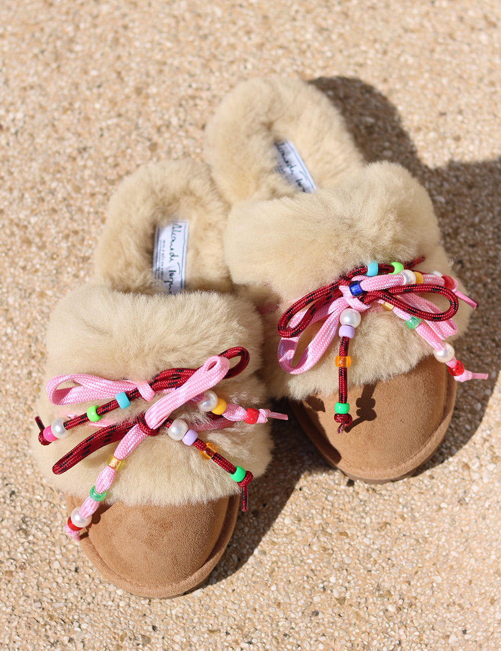 Carrie Pink shearling mules flat lay — pair of cream shearling mules with handcrafted multicolour beaded cord detailing in pink and bordeaux on caramel suede, styled overhead on sandy ground, handcrafted in Portugal by Alameda Turquesa