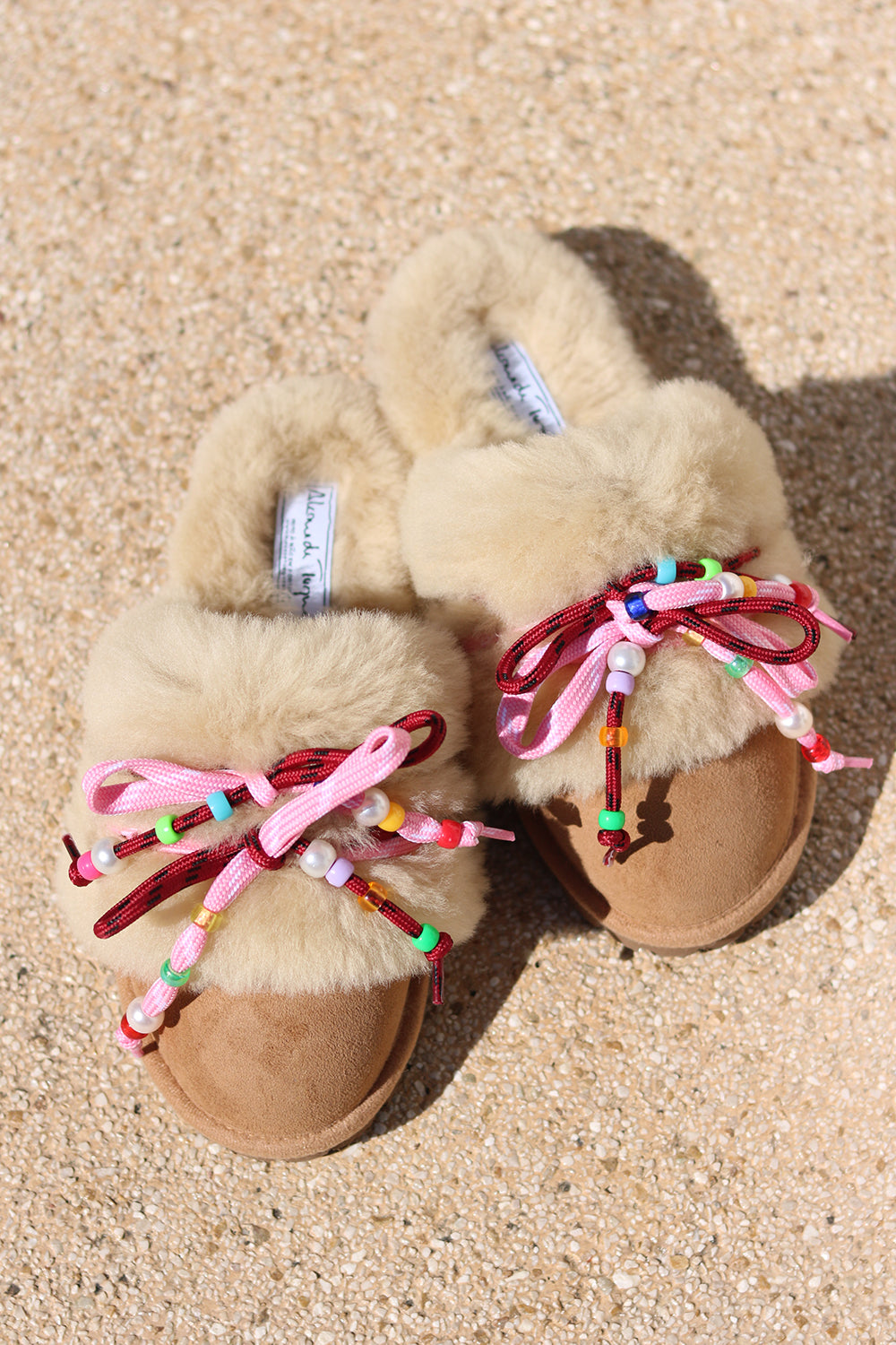 Carrie Pink shearling mules flat lay — pair of cream shearling mules with handcrafted multicolour beaded cord detailing in pink and bordeaux on caramel suede, styled overhead on sandy ground, handcrafted in Portugal by Alameda Turquesa