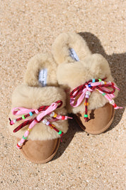 Carrie Pink shearling mules flat lay — pair of cream shearling mules with handcrafted multicolour beaded cord detailing in pink and bordeaux on caramel suede, styled overhead on sandy ground, handcrafted in Portugal by Alameda Turquesa