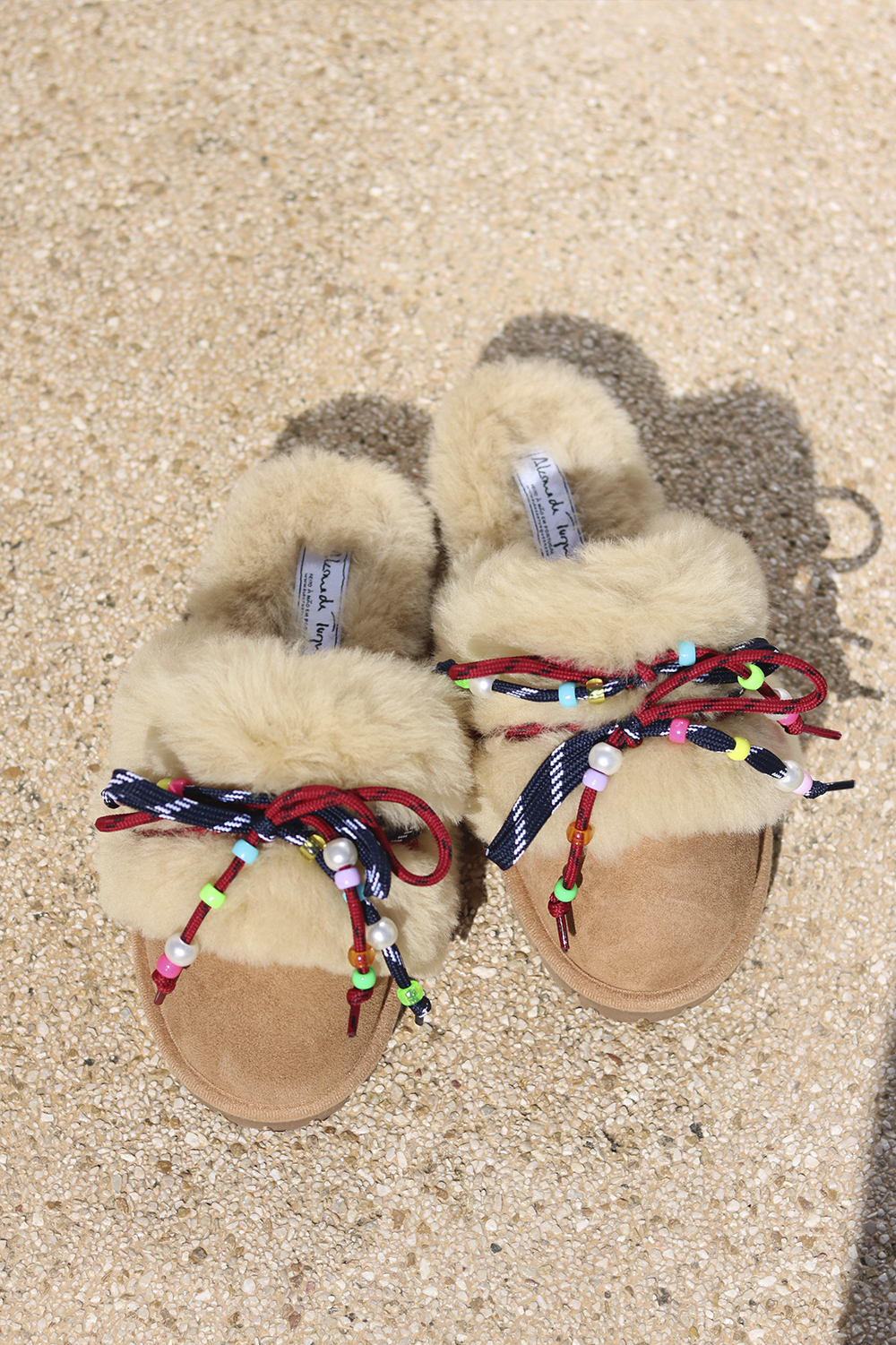 Carrie Navy shearling mules flat lay — pair of cream shearling mules with handcrafted multicolour beaded cord detailing in navy and red on caramel suede, styled overhead on sandy ground, handcrafted in Portugal by Alameda Turquesa
