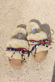 Carrie Navy shearling mules flat lay — pair of cream shearling mules with handcrafted multicolour beaded cord detailing in navy and red on caramel suede, styled overhead on sandy ground, handcrafted in Portugal by Alameda Turquesa