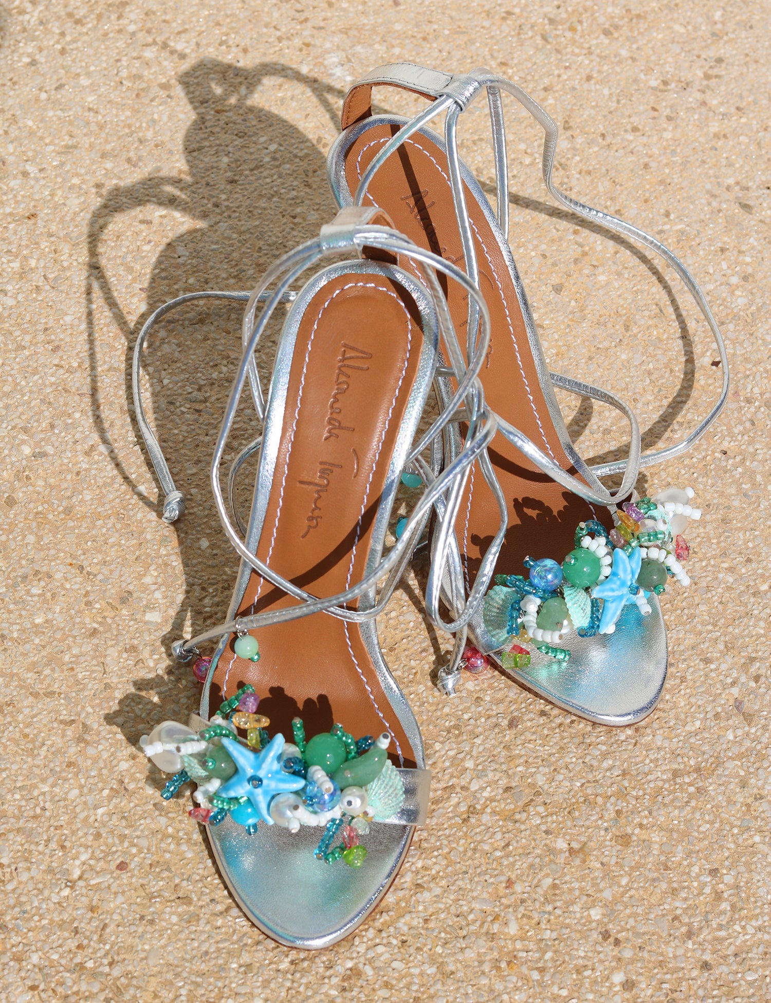 Aquamarine heels flat lay — silver stiletto sandals with wraparound ankle laces, tan leather insole with Alameda Turquesa branding, and handcrafted aquamarine, turquoise, green and coral starfish sea-motif embellishments at the toe handcrafted in Portugal by Alameda Turquesa