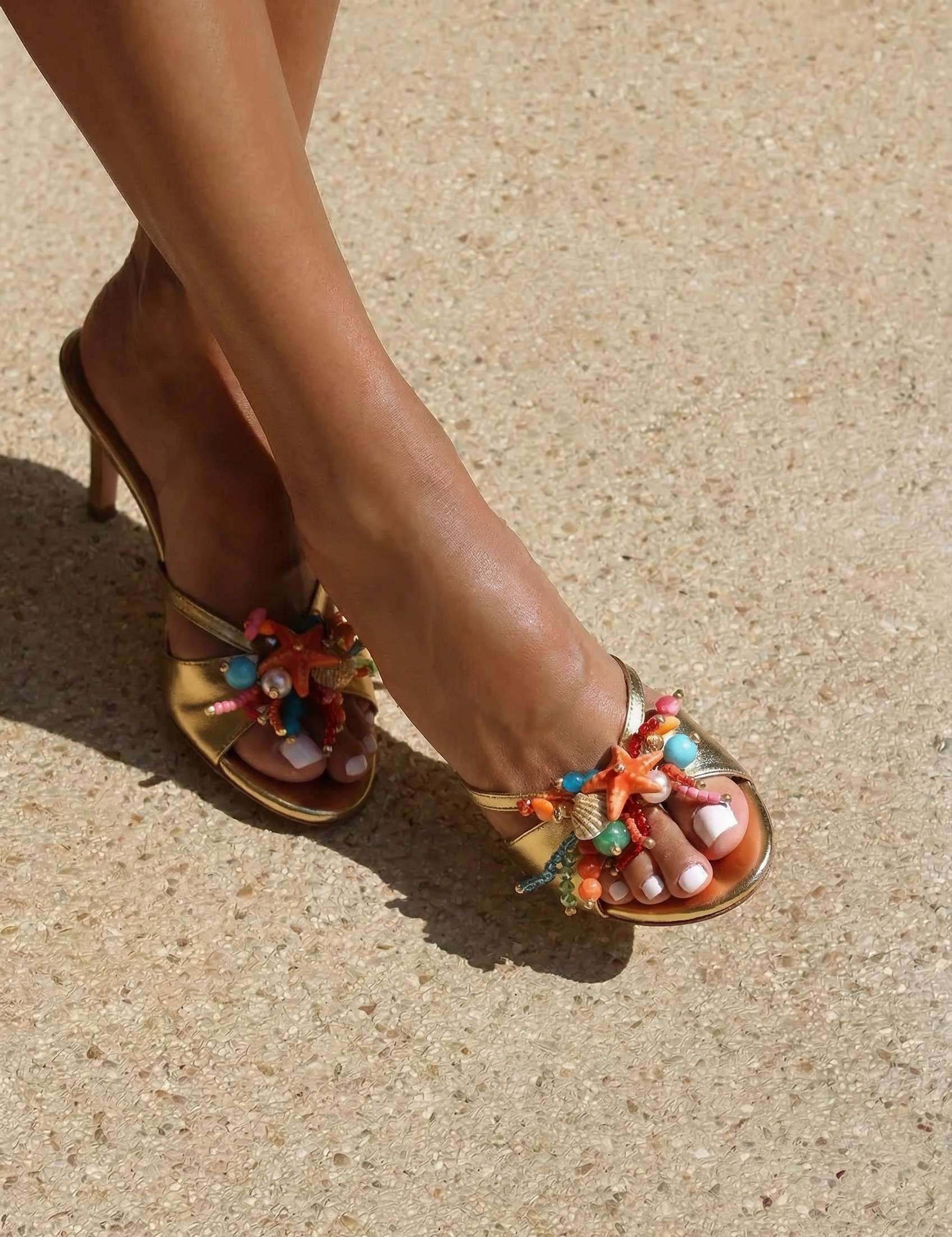 Bahamas mules side profile on foot — gold leather mid-heel open-toe mule with hand-painted starfish, natural shells, pearl and multicolor bead embellishments in coral, turquoise and pink, shot in sunlight on sand, handcrafted in Portugal by Alameda Turquesa