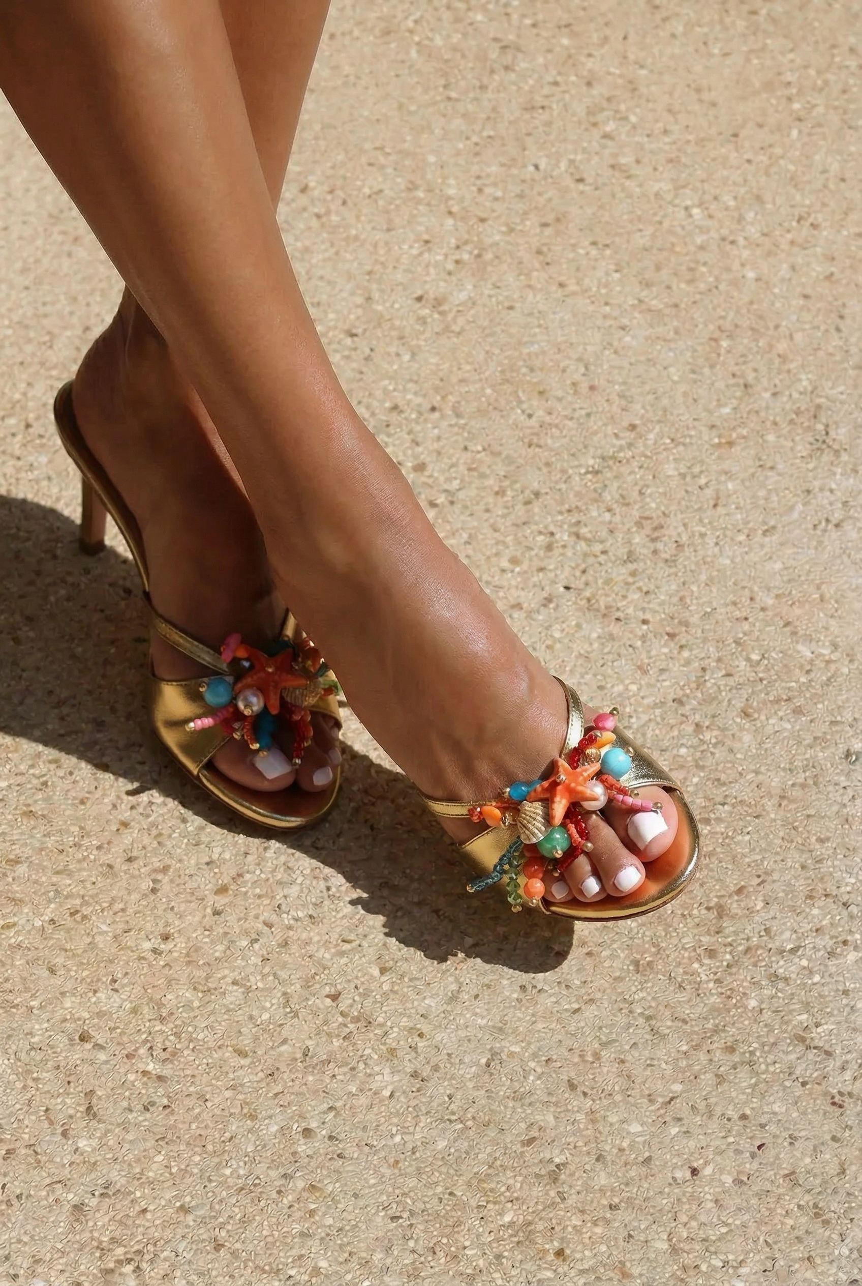 Bahamas mules side profile on foot — gold leather mid-heel open-toe mule with hand-painted starfish, natural shells, pearl and multicolor bead embellishments in coral, turquoise and pink, shot in sunlight on sand, handcrafted in Portugal by Alameda Turquesa