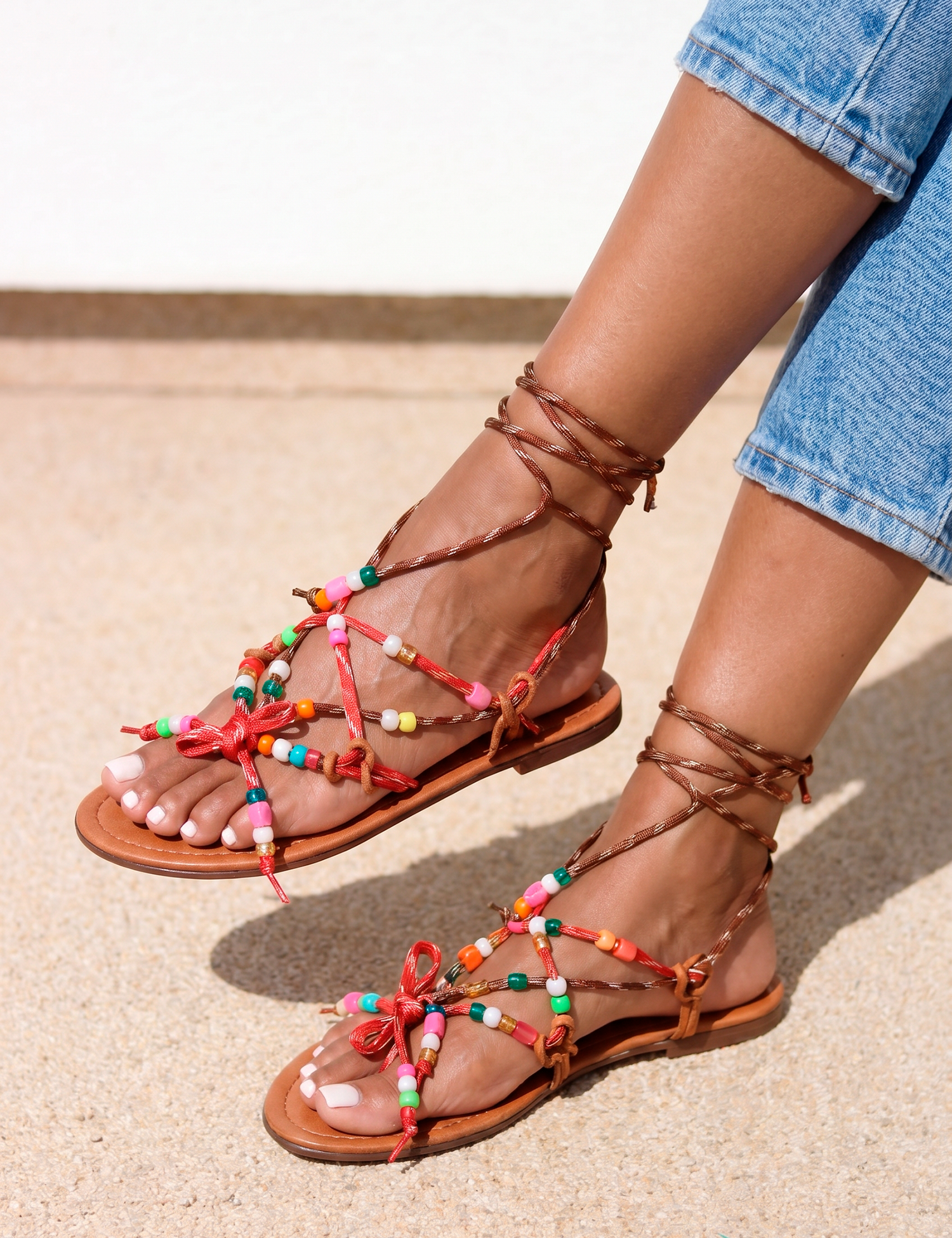 Lilo sandals pair view — tan braided leather flat sandals with multicolour handcrafted beaded straps, red bow details and lace-up ankle ties, worn on body with jeans, handcrafted in Portugal by Alameda Turquesa