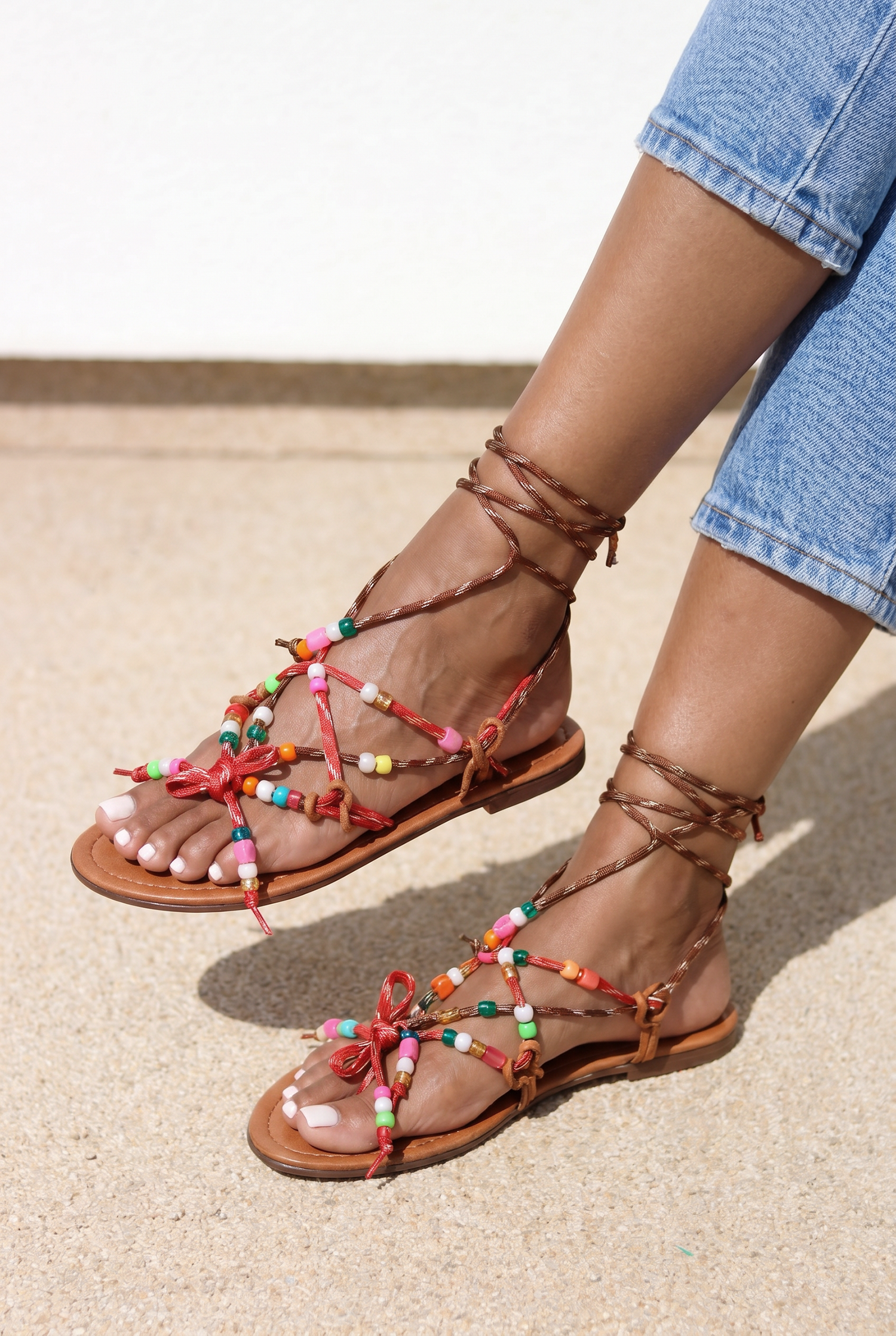 Lilo sandals pair view — tan braided leather flat sandals with multicolour handcrafted beaded straps, red bow details and lace-up ankle ties, worn on body with jeans, handcrafted in Portugal by Alameda Turquesa