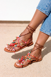 Lilo sandals pair view — tan braided leather flat sandals with multicolour handcrafted beaded straps, red bow details and lace-up ankle ties, worn on body with jeans, handcrafted in Portugal by Alameda Turquesa