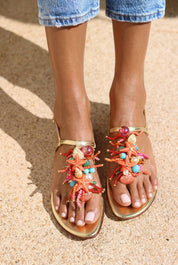 Atlântica sandals front view on both feet — gold leather toe-post flat sandals with hand-applied coral starfish, natural shells, pearls, turquoise stones and multicolour bead embellishments, styled with denim shorts in sunlight on sand, handcrafted in Portugal by Alameda Turquesa