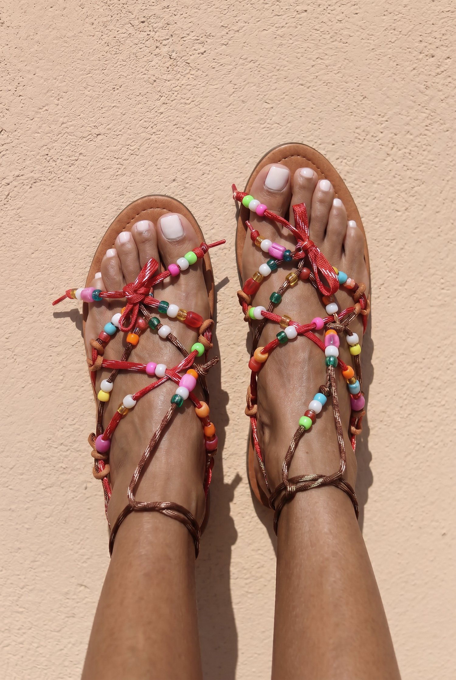Lilo sandals top view — tan braided leather flat sandals with multicolour handcrafted beaded straps and red bow details, worn on body against a terracotta wall, handcrafted in Portugal by Alameda Turquesa