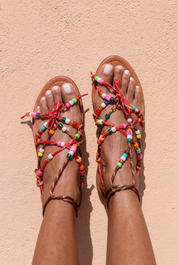 Lilo sandals top view — tan braided leather flat sandals with multicolour handcrafted beaded straps and red bow details, worn on body against a terracotta wall, handcrafted in Portugal by Alameda Turquesa