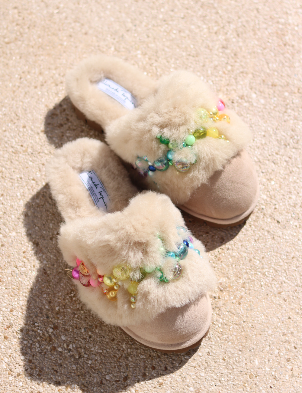 Pinctada Vanilla Rainbow shearling mules flat lay — pair of ivory shearling mules with handcrafted rainbow pearl embellishments in pink, orange, yellow, green and aqua on sand suede, styled overhead on sandy ground, handcrafted in Portugal by Alameda Turquesa