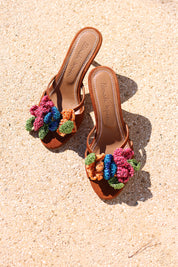 Botanica mules flat lay on sand — tan leather flat mules with handcrafted crochet flower embellishments in pink, blue, orange and green, Alameda Turquesa leather insole, handcrafted in Portugal by Alameda Turquesa