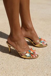 Bahamas mules front view on both feet — gold leather stiletto heel open-toe mules with hand-painted starfish, natural shells, pearl and multicolour bead embellishments in coral, turquoise and pink, shot in sunlight on sand, handcrafted in Portugal by Alameda Turquesa
