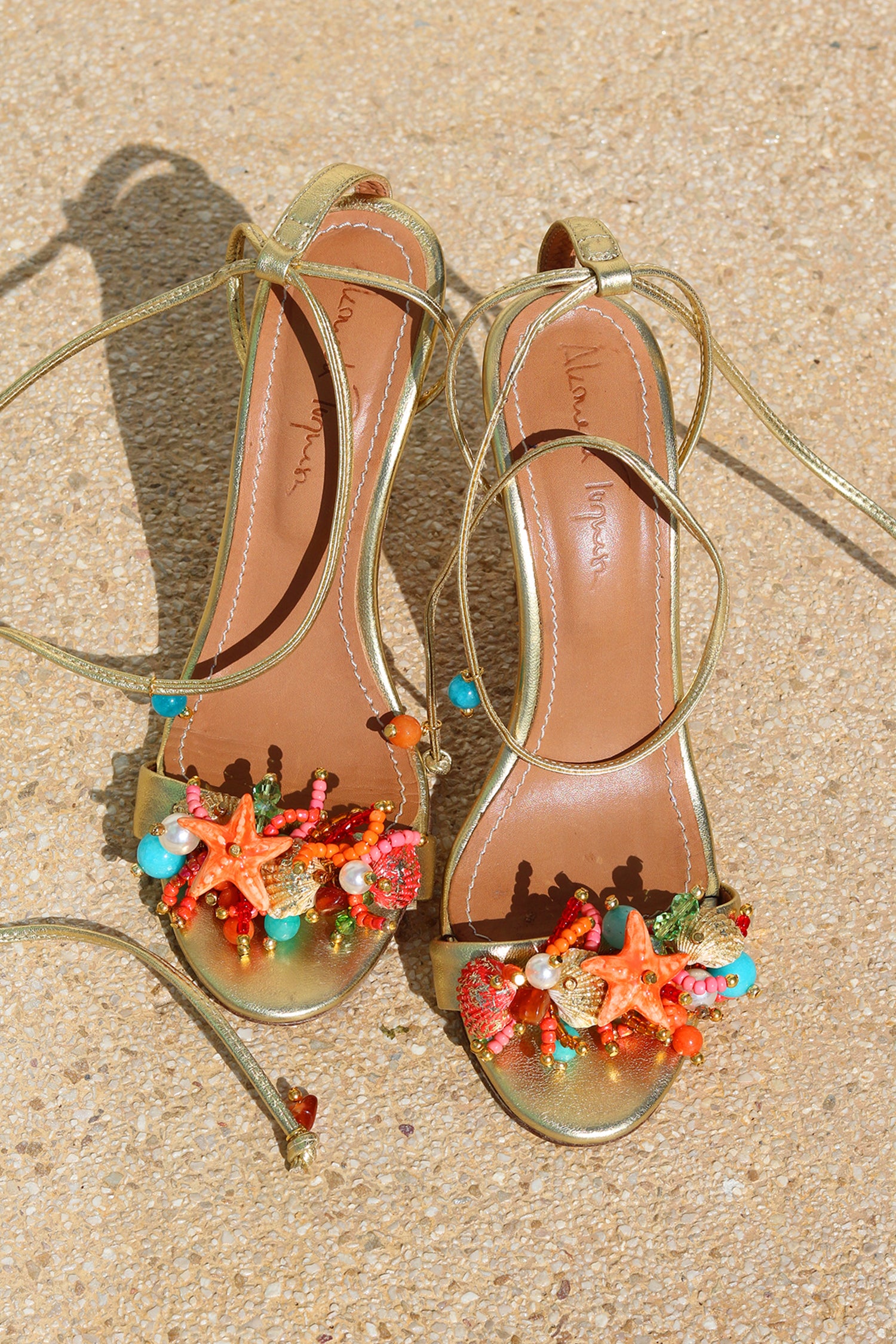Bahamas heels flat lay — gold stiletto sandals with wraparound ankle laces, tan leather insole with Alameda Turquesa branding, and handcrafted coral, starfish, turquoise and pearl sea-motif embellishments at the toe, shot on sand, handcrafted in Portugal by Alameda Turquesa