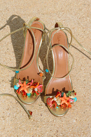 Bahamas heels flat lay — gold stiletto sandals with wraparound ankle laces, tan leather insole with Alameda Turquesa branding, and handcrafted coral, starfish, turquoise and pearl sea-motif embellishments at the toe, shot on sand, handcrafted in Portugal by Alameda Turquesa
