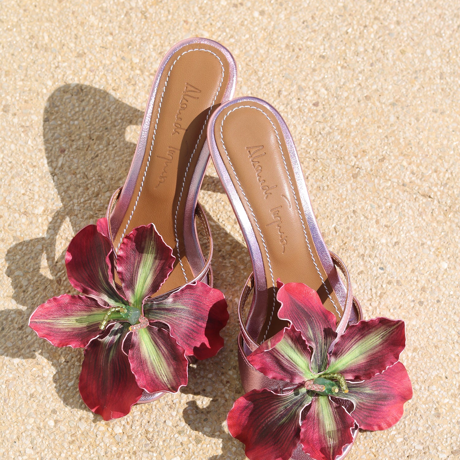 La Isla Rosaire mules flat lay — pink metallic leather open-toe mules with oversized handcrafted deep crimson orchid appliqué, shot on sand, handcrafted in Portugal by Alameda Turquesa