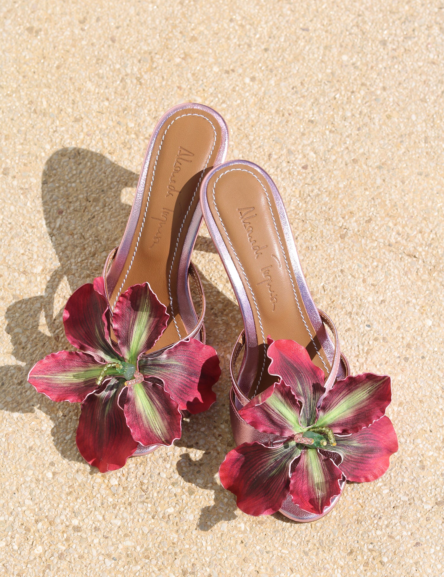 La Isla Rosaire mules flat lay — pink metallic leather open-toe mules with oversized handcrafted deep crimson orchid appliqué, shot on sand, handcrafted in Portugal by Alameda Turquesa