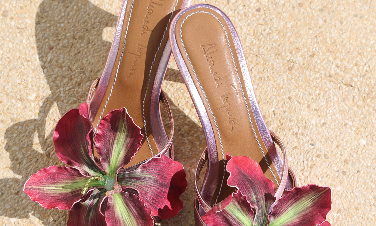 La Isla Rosaire mules flat lay — pink metallic leather open-toe mules with oversized handcrafted deep crimson orchid appliqué, shot on sand, handcrafted in Portugal by Alameda Turquesa