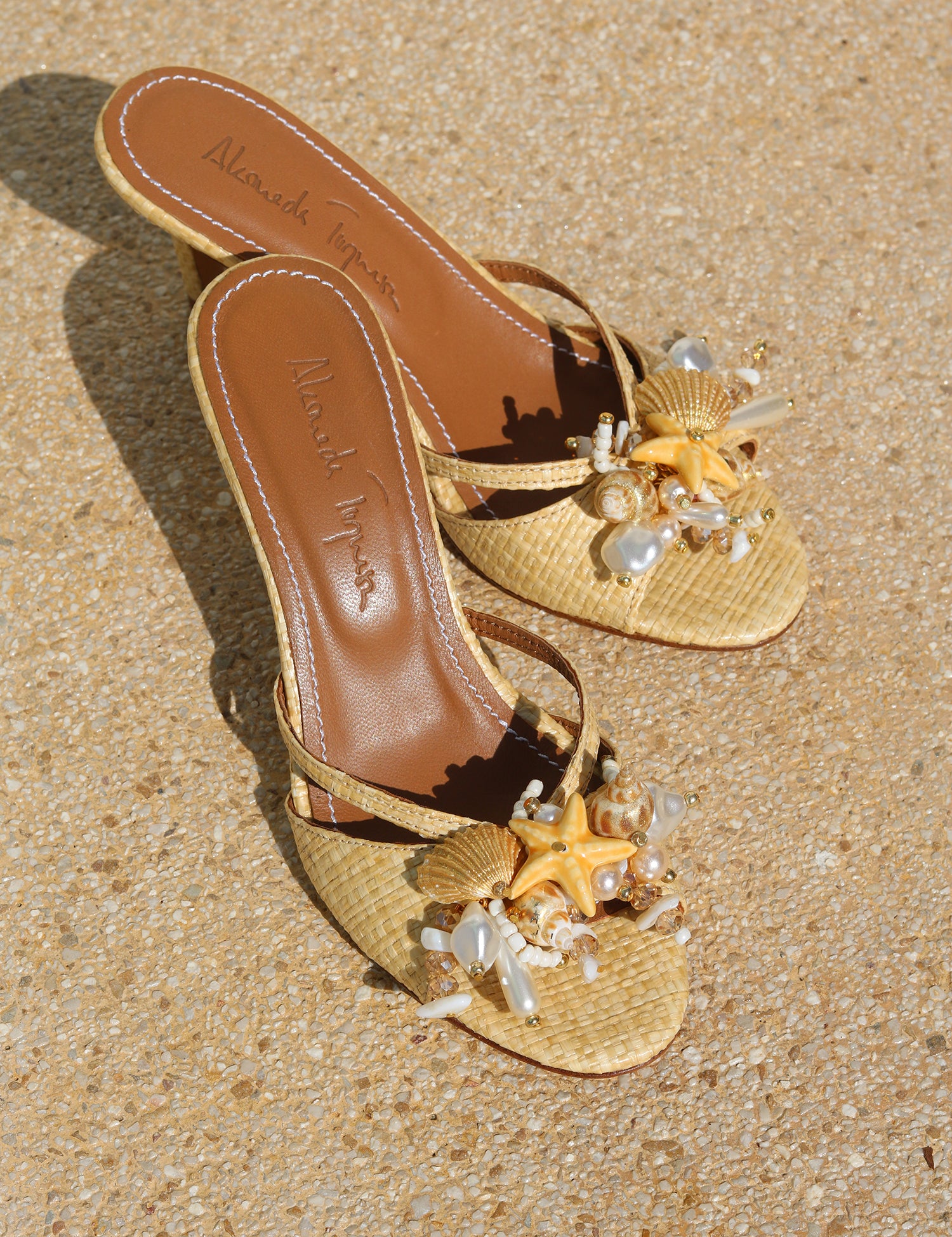 La Isla Miramar mules flat lay — natural raffia open-toe mules with hand-applied shell, pearl and starfish embellishments, Alameda Turquesa insole visible, shot on sand, handcrafted in Portugal by Alameda Turquesa