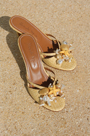 La Isla Miramar mules flat lay — natural raffia open-toe mules with hand-applied shell, pearl and starfish embellishments, Alameda Turquesa insole visible, shot on sand, handcrafted in Portugal by Alameda Turquesa