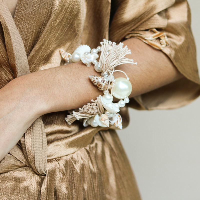 Ariel bracelet on wrist — handcrafted pearl, seashell and natural tassel bracelet with oversized pearl centrepiece, worn against gold fabric, handcrafted in Portugal by Alameda Turquesa