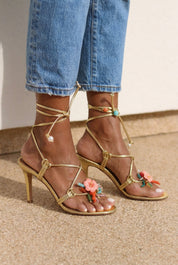 Coralie sandals front view both feet — gold leather multi-strap wrap stiletto heels with pearl and turquoise bead ankle detail and hand-painted ceramic hibiscus embellishments, styled with denim jeans, handcrafted in Portugal by Alameda Turquesa