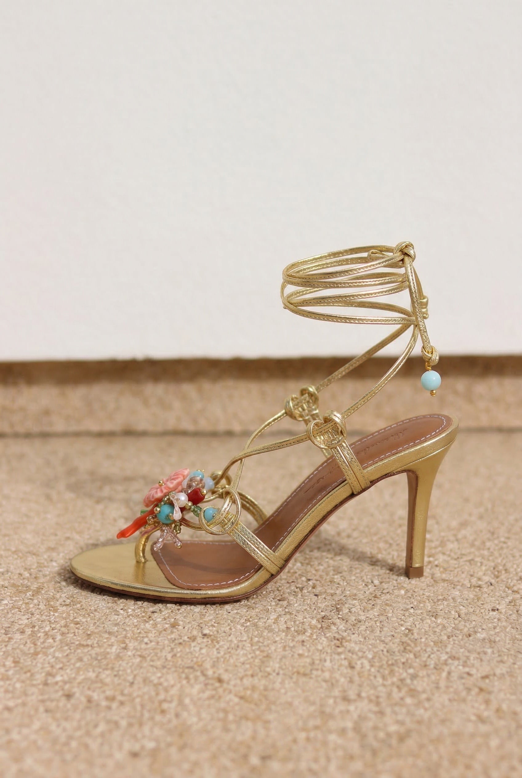 Coralie sandal single shoe side profile — gold leather multi-strap wrap stiletto heel with hand-painted ceramic hibiscus and turquoise and coral bead embellishments, against pale wall on sand, handcrafted in Portugal by Alameda Turquesa